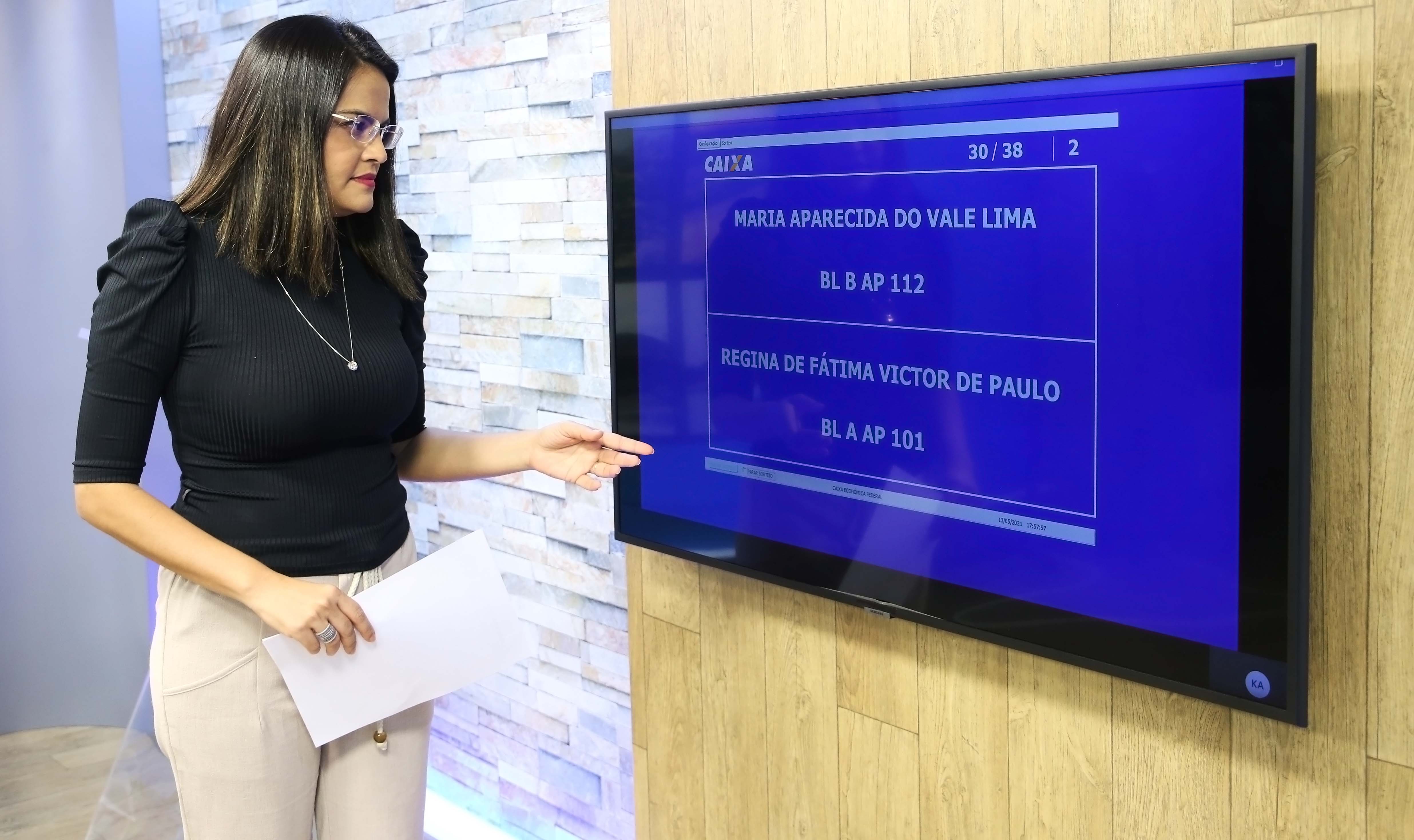 mulher na frente de uma tela de TV na qual aparecem os nomes das pessoas sorteadas 